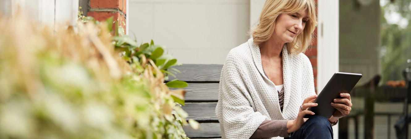 A mature woman sitting outside her home while using a tablet.
