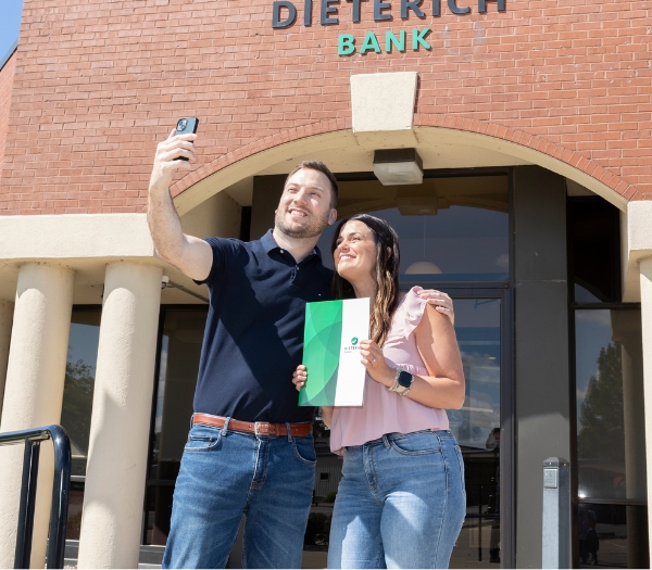 A husband holds up his phone to take a selfie with his wife holding their new account outside of a bank.