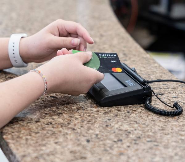 A hand guides a debit card towards a terminal sitting on a desk.