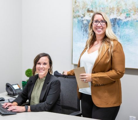 One woman smiles as she types on her computer at her desk as another woman smiles standing behind her.