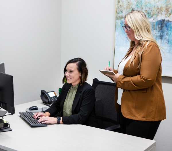 One woman types on her computer at her desk as another woman stands behind her writing on a notepad.
