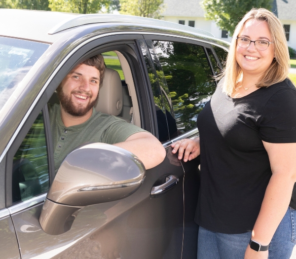 A man smiles sitting in the driver's seat of a new car as his wife smiles standing by the car.