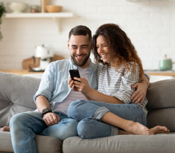 A young woman and her husband sits on a couch while using her phone.