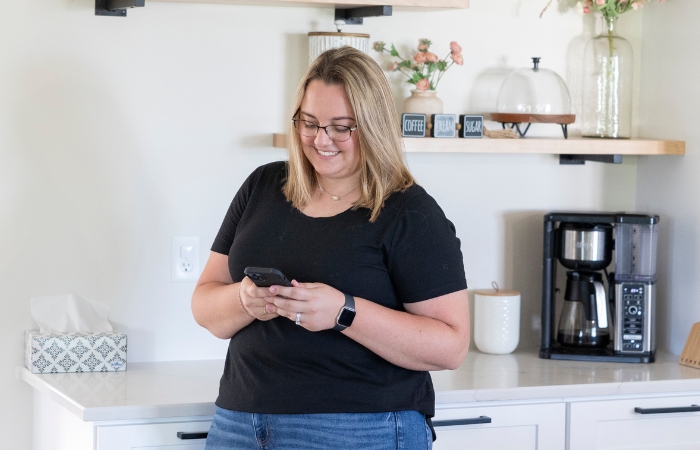 A woman smiles while looking at her phone while standing in a kitchen.