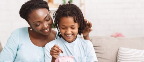 A child sits on their mother's lap while dropping a penny in a piggy bank.