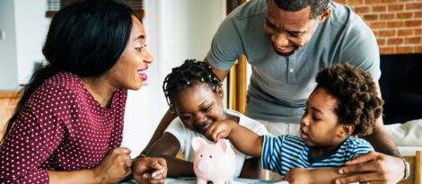 A Black family sits around a pink piggy bank, with one child placing a coin in the bank.