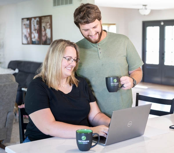 A woman sits at kitchen table typing on her computer as her husband stands by her holding a coffee mug.