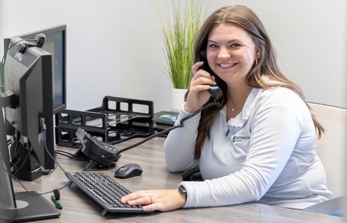 A young female bank employee looks at the camera smiling while talking on the phone.