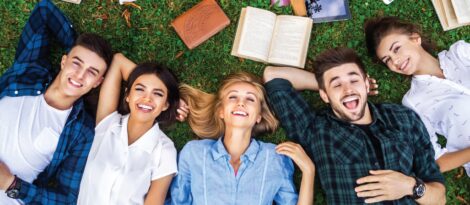 A group of smiling young adults lay on a lawn surrounded by books.