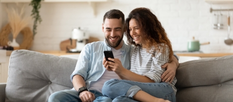 A young woman and her husband sits on a couch while using her phone.