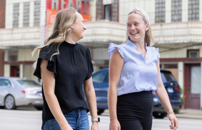 Two women smile at each other as they walk on a sidewalk together.