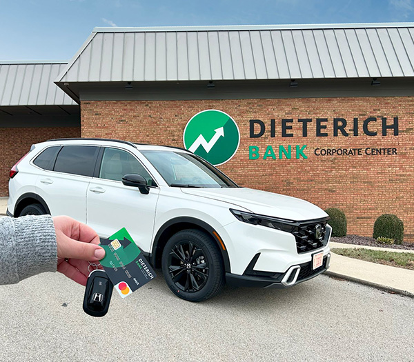 A white Honda CRV is parked in front of the Dieterich Bank Corporate Office with a woman's hands holding a key fob with a Dieterich Bank debit card attached to the key ring in front of the car.