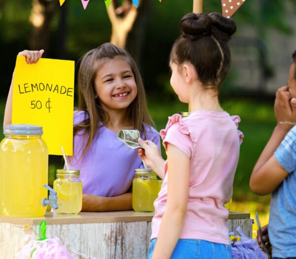 1 young girl is selling lemonade for 50 cents. Another young girl is holding out a dollar to pay for a cup of lemonade.
